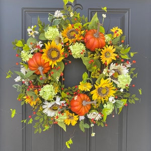 May include: A vibrant autumn wreath featuring sunflowers, orange pumpkins, and green foliage. The wreath is adorned with yellow and white flowers, red berries, and green hydrangeas. This decorative wreath is perfect for fall and seasonal home decor.