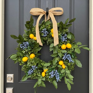 May include: A wreath made of green leaves and blue berries with yellow lemons hanging from it. The wreath is tied with a brown burlap ribbon and is hanging on a black door.