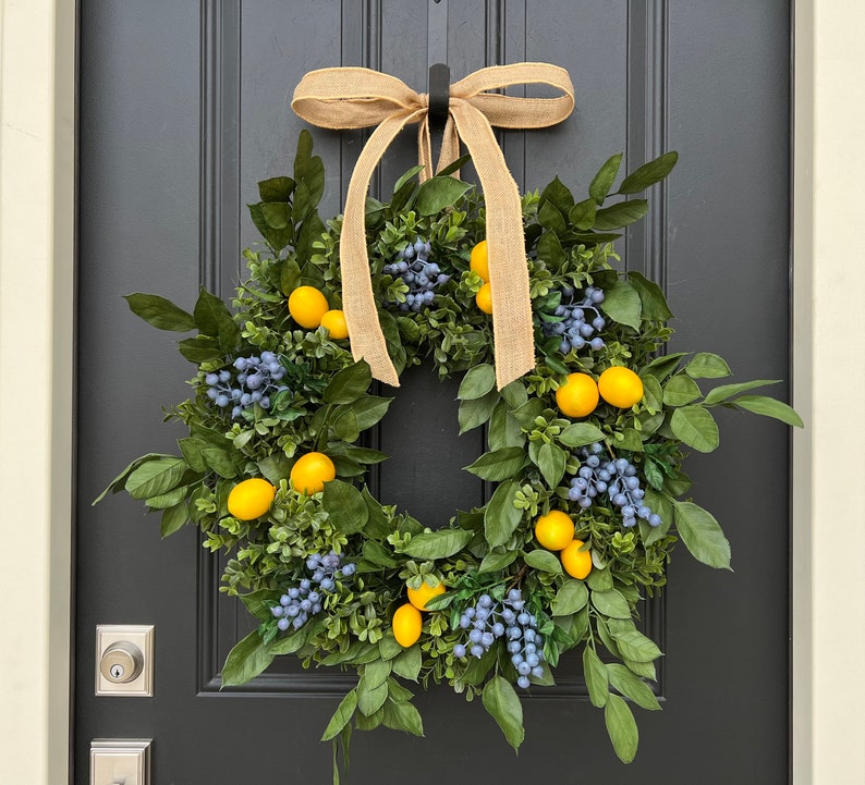 May include: A wreath made of green leaves and blue berries with yellow lemons hanging from it. The wreath is tied with a brown burlap ribbon and is hanging on a black door.