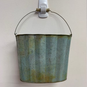 May include: A weathered, galvanized metal bucket with a ribbed design and a wire handle. The bucket has a blue-green patina and is hanging from a white wall hook. The bucket is empty.
