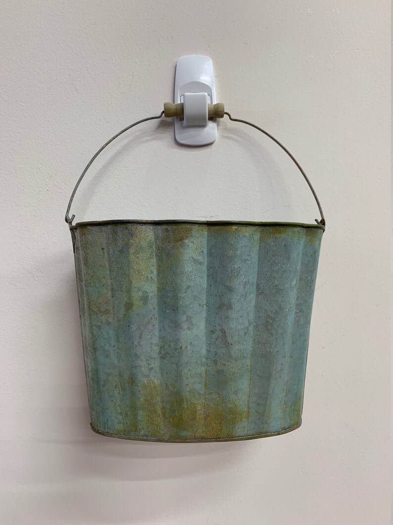 May include: A weathered, galvanized metal bucket with a ribbed design and a wire handle. The bucket has a blue-green patina and is hanging from a white wall hook. The bucket is empty.