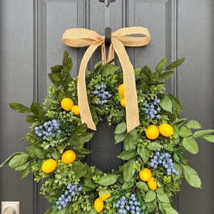 May include: A green wreath with yellow lemons and blue berries, decorated with a brown burlap bow, hanging on a black door.