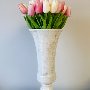 May include: A white, distressed ceramic vase filled with pink and white tulips. The vase has a pedestal base and a flared top. The tulips have green stems and leaves, creating a fresh, spring-like arrangement.