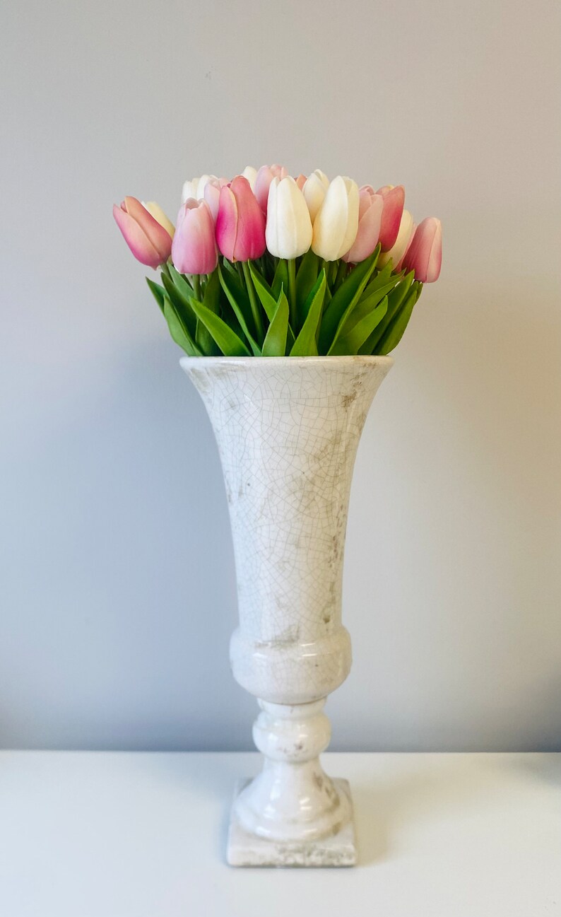 May include: A white, distressed ceramic vase filled with pink and white tulips. The vase has a pedestal base and a flared top. The tulips have green stems and leaves, creating a fresh, spring-like arrangement.