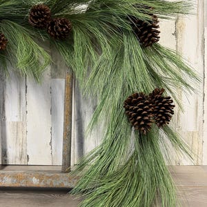 May include: A decorative garland featuring long, green pine needles and several brown pine cones. The garland is arranged in a flowing, naturalistic style, perfect for holiday or seasonal decor. The background is a rustic, weathered wood.