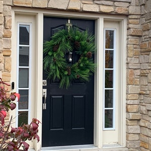 May include: A black front door with a green pine wreath adorned with pinecones and a small lantern. The door is flanked by sidelight windows with white trim. The house exterior features a stone facade and a red bush.