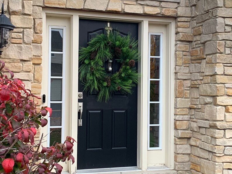 May include: A black front door with a green pine wreath adorned with pinecones and a small lantern. The door is flanked by sidelight windows with white trim. The house exterior features a stone facade and a red bush.