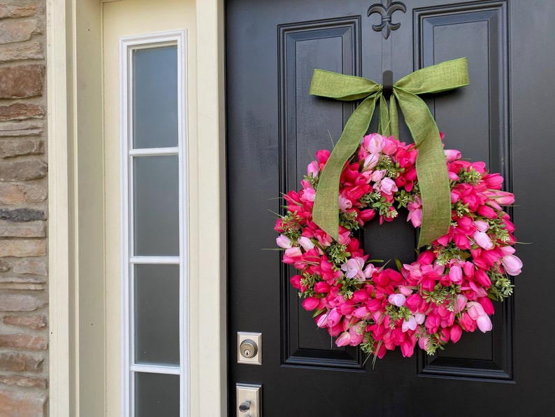 May include: A pink and green tulip wreath with a green burlap bow hanging on a black door.