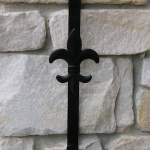 May include: Black metal wreath hanger with a fleur-de-lis design. The hanger is mounted on a stone wall.