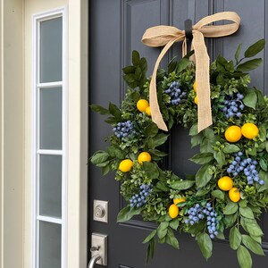 May include: A lemon and blueberry wreath with a burlap bow hanging on a black door. The wreath is made of green leaves, yellow lemons, and blue berries.