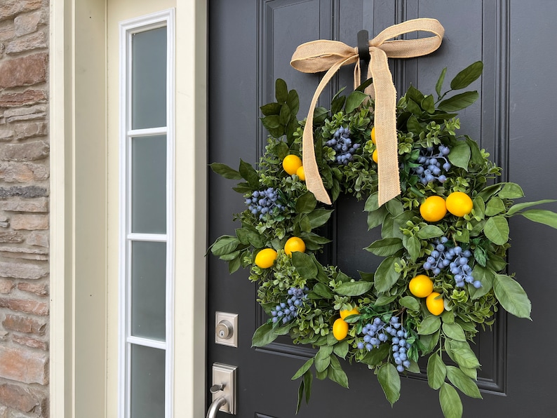 May include: A lemon and blueberry wreath with a burlap bow hanging on a black door. The wreath is made of green leaves, yellow lemons, and blue berries.
