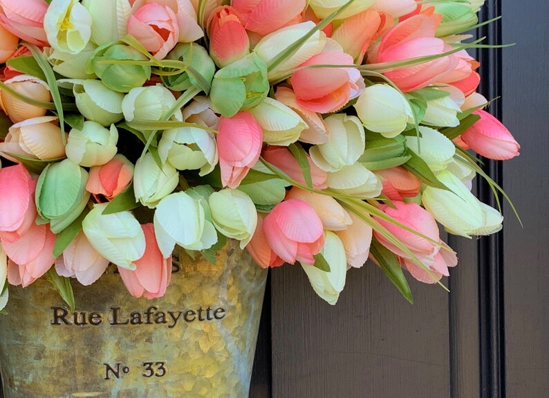 May include: A floral arrangement of artificial tulips in shades of pink, peach, and white, with green leaves. The flowers are in a galvanized metal bucket with the text "Rue Lafayette No. 33".