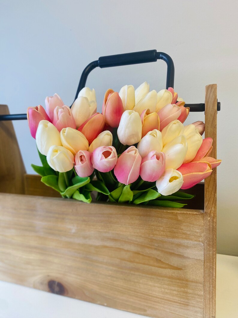 May include: A wooden box with a black metal handle filled with a bouquet of colorful tulips. The tulips are in shades of pink, peach, and white, with green leaves. The box has a natural wood finish.