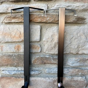 May include: Two metal over-the-door hooks. One is black, and the other is bronze. Each hook has a rectangular vertical bar with a horizontal bar at the top and a curved hook at the bottom. The hooks are designed to hang over a door.