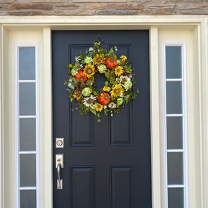 May include: A fall-themed wreath with sunflowers, pumpkins, and greenery hangs on a black front door. The door is framed by cream-colored trim and sidelights, set against a stone exterior.