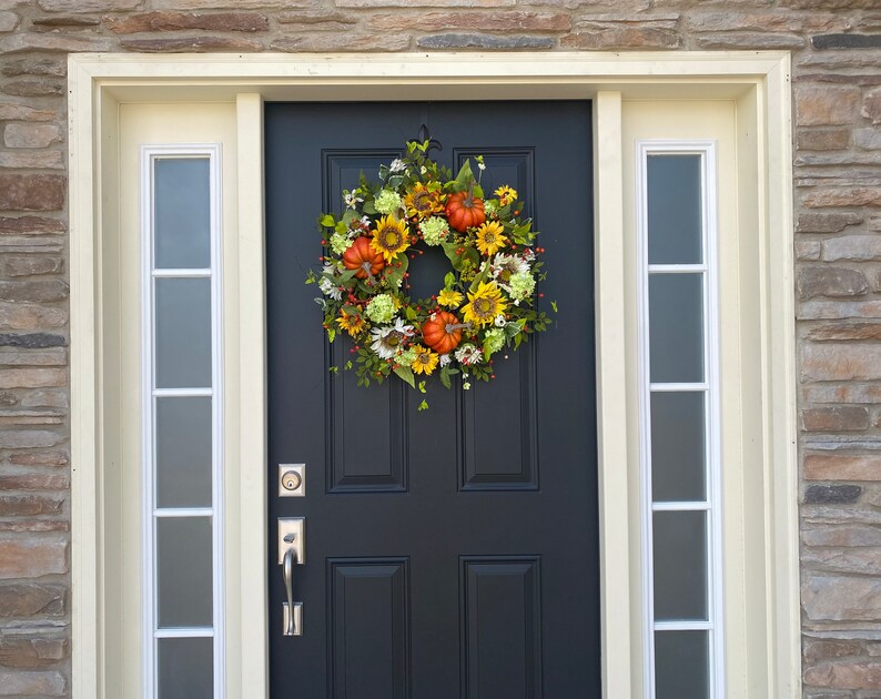May include: A fall-themed wreath with sunflowers, pumpkins, and greenery hangs on a black front door. The door is framed by cream-colored trim and sidelights, set against a stone exterior.