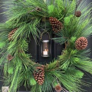 May include: A festive Christmas wreath with lush green pine needles and brown pine cones. A small metal lantern with a lit candle hangs in the center. The word "JOY" is displayed above the wreath.