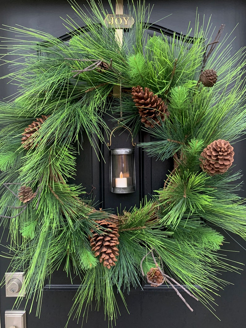 May include: A festive Christmas wreath with lush green pine needles and brown pine cones. A small metal lantern with a lit candle hangs in the center. The word "JOY" is displayed above the wreath.