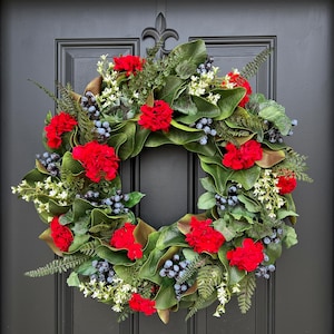 May include: A vibrant, circular wreath with a mix of red flowers, green leaves, and small blue berries. The wreath is adorned with white accents and fern-like foliage, creating a full and textured appearance. It is displayed on a black door.