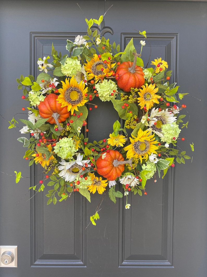 May include: A vibrant autumn wreath featuring sunflowers, orange pumpkins, and green foliage. The wreath is adorned with yellow and white flowers, red berries, and green hydrangeas. This decorative wreath is perfect for fall and seasonal home decor.