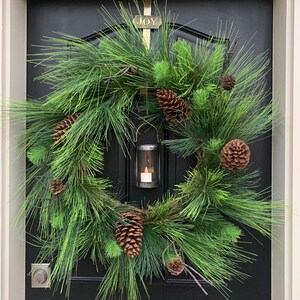 May include: A festive evergreen wreath with faux pine needles and brown pine cones. A small lantern with a lit candle is centered within the wreath. The wreath hangs on a black door, with a gold sign above that reads "JOY".