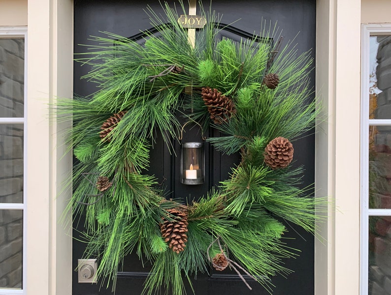 May include: A festive evergreen wreath with faux pine needles and brown pine cones. A small lantern with a lit candle is centered within the wreath. The wreath hangs on a black door, with a gold sign above that reads "JOY".