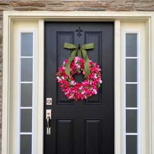 May include: A black front door with a pink and green floral wreath. The wreath is made of artificial flowers and has a green ribbon bow.