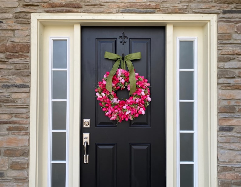 May include: A black front door with a pink and green floral wreath. The wreath is made of artificial flowers and has a green ribbon bow.