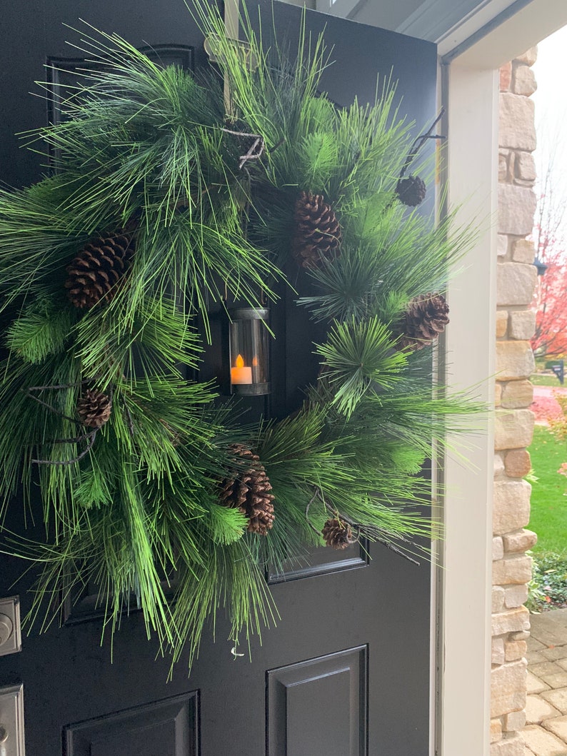May include: A green pine wreath with several brown pine cones and a small lantern with a flickering candle is hanging on a black door. The wreath is a full, circular shape, and the pine needles are a vibrant green.