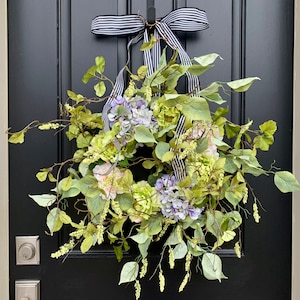 May include: A decorative wreath featuring green and purple faux flowers and foliage. The wreath is adorned with a black and white striped ribbon and is hanging on a black door. The wreath is a beautiful accent for any home.