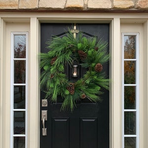 May include: A black front door with a decorative evergreen wreath, adorned with pinecones and a small lantern. The word "JOY" is above the door. The door is framed by cream-colored trim and sidelights, set against a stone exterior.