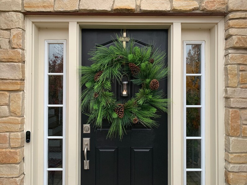 May include: A black front door with a decorative evergreen wreath, adorned with pinecones and a small lantern. The word "JOY" is above the door. The door is framed by cream-colored trim and sidelights, set against a stone exterior.