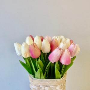 May include: A bouquet of tulips in a woven basket. The tulips are a mix of white, light pink, and deep pink. The basket is made of light-colored woven material. Green leaves are visible.