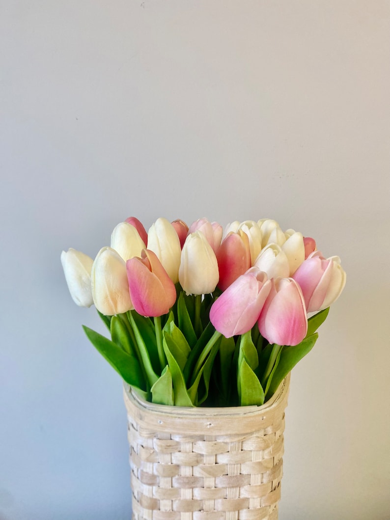 May include: A bouquet of tulips in a woven basket. The tulips are a mix of white, light pink, and deep pink. The basket is made of light-colored woven material. Green leaves are visible.