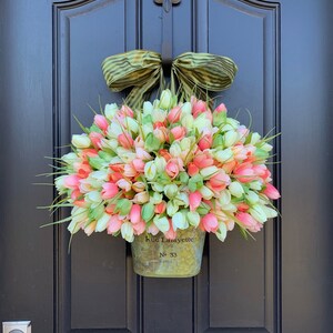 May include: A spring wreath featuring a metal bucket overflowing with pink, white, and coral tulip buds. The bucket is inscribed with "Rue Lafayette" and "No. 33". A green and brown striped bow tops the arrangement, hanging on a black door.
