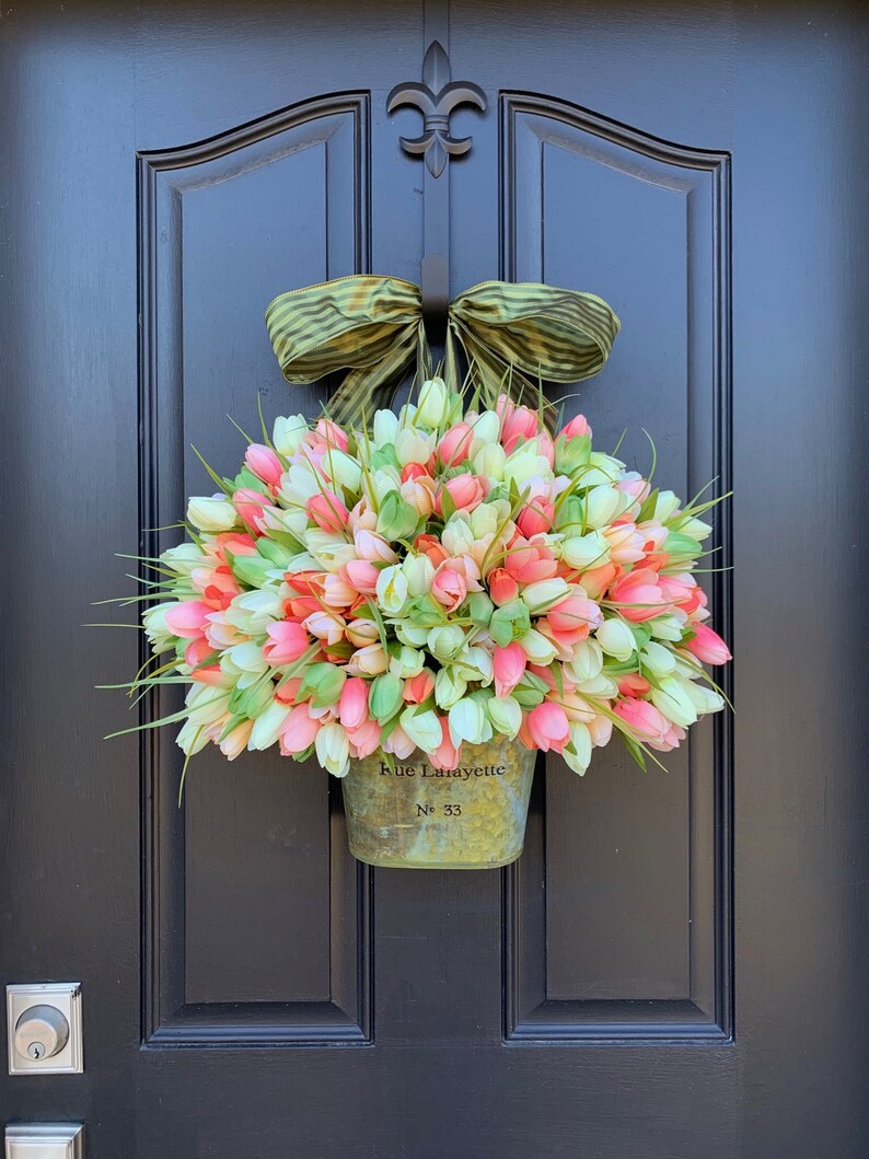 May include: A spring wreath featuring a metal bucket overflowing with pink, white, and coral tulip buds. The bucket is inscribed with "Rue Lafayette" and "No. 33". A green and brown striped bow tops the arrangement, hanging on a black door.