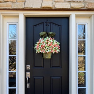 May include: A black front door with a floral wreath. The wreath is made of pink, white, and green tulips in a decorative metal container. The door is framed by white trim and stone siding.