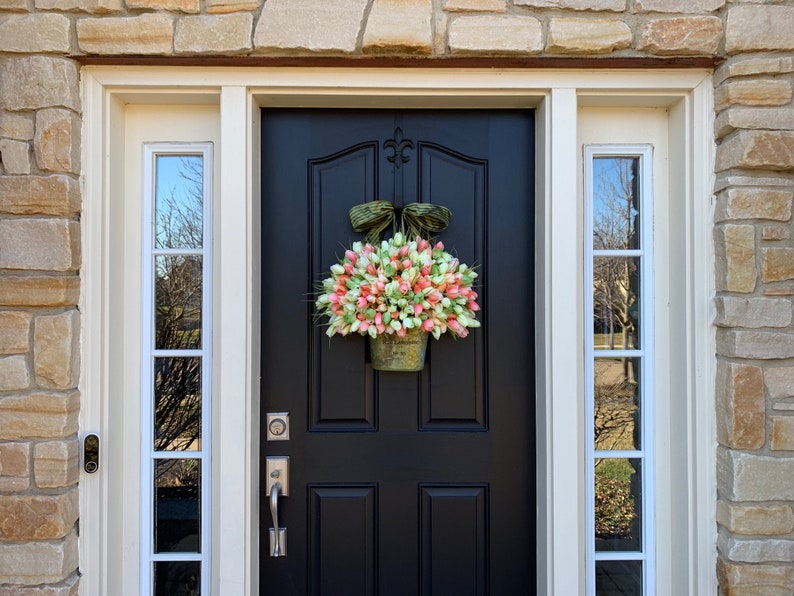 May include: A black front door with a floral wreath. The wreath is made of pink, white, and green tulips in a decorative metal container. The door is framed by white trim and stone siding.