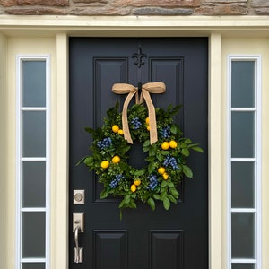 May include: A black front door with a wreath made of green leaves, yellow lemons, and blue berries. The wreath is tied with a brown burlap ribbon.