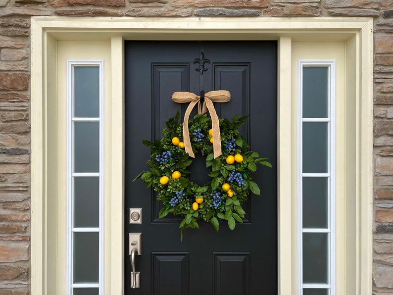 May include: A black front door with a wreath made of green leaves, yellow lemons, and blue berries. The wreath is tied with a brown burlap ribbon.