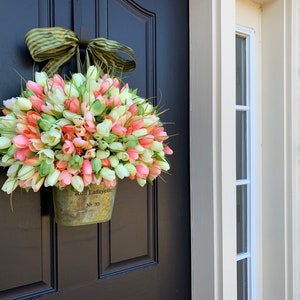 May include: A tulip wreath in a metal bucket hangs on a black door. The wreath features pink, white, and green tulips, accented by green grass. A green and brown striped bow is at the top. The bucket has the text "Rue Lafayette No 33".
