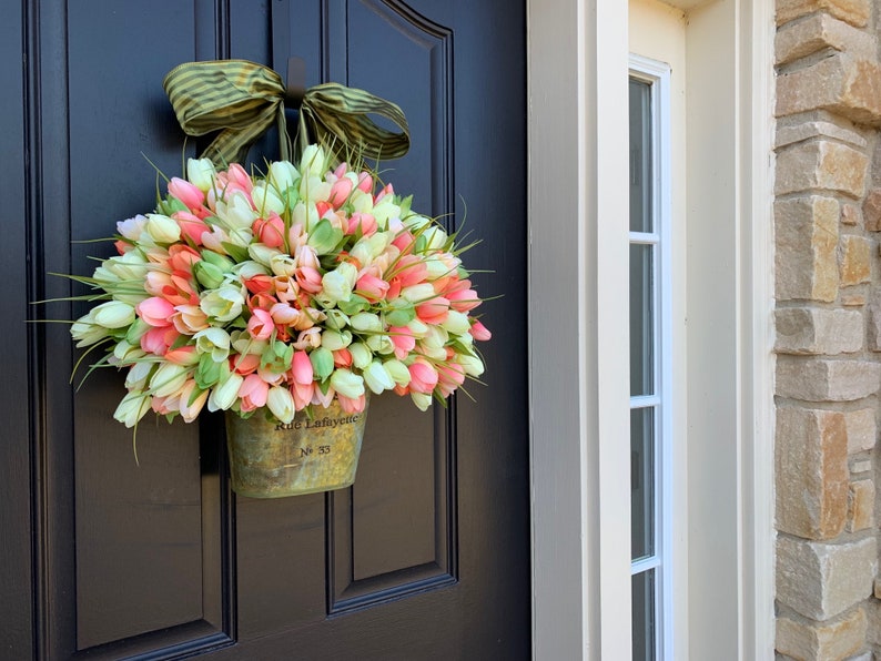 May include: A tulip wreath in a metal bucket hangs on a black door. The wreath features pink, white, and green tulips, accented by green grass. A green and brown striped bow is at the top. The bucket has the text "Rue Lafayette No 33".