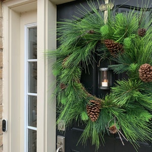 May include: A festive Christmas wreath with lush green pine needles and several brown pine cones. A small, silver lantern with a lit candle hangs from the wreath. The wreath is on a black door with a gold "JOY" plaque.
