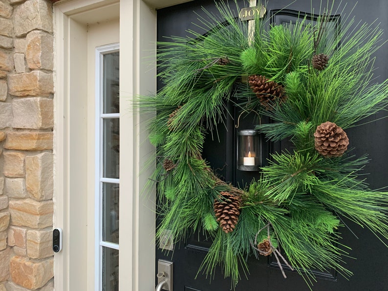 May include: A festive Christmas wreath with lush green pine needles and several brown pine cones. A small, silver lantern with a lit candle hangs from the wreath. The wreath is on a black door with a gold "JOY" plaque.