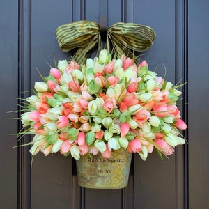 May include: A spring-themed door wreath featuring a galvanized metal container overflowing with colorful tulips in shades of pink, white, and coral. A green and tan striped bow is attached at the top. The container is labeled "Rue Lafayette" and "No. 33".
