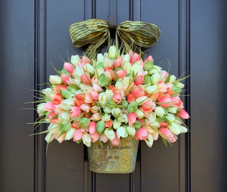 May include: A spring-themed door wreath featuring a galvanized metal container overflowing with colorful tulips in shades of pink, white, and coral. A green and tan striped bow is attached at the top. The container is labeled "Rue Lafayette" and "No. 33".