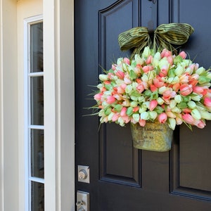 May include: A spring-themed wreath of pink, peach, and white tulips in a galvanized metal bucket, hanging on a black door. The bucket is labeled "Rue Lafayette No. 33" and is topped with a green and tan striped bow.