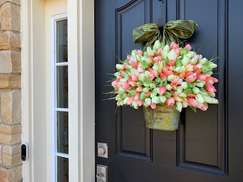 May include: A spring-themed wreath of pink, peach, and white tulips in a galvanized metal bucket, hanging on a black door. The bucket is labeled "Rue Lafayette No. 33" and is topped with a green and tan striped bow.