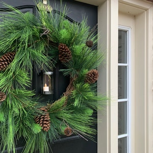 May include: A festive evergreen wreath with pinecones and a small candle lantern. The wreath is displayed on a black door, with a glimpse of a window and stone wall in the background. The wreath is a classic holiday decoration.