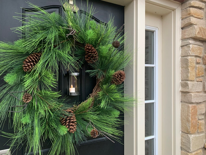 May include: A festive evergreen wreath with pinecones and a small candle lantern. The wreath is displayed on a black door, with a glimpse of a window and stone wall in the background. The wreath is a classic holiday decoration.